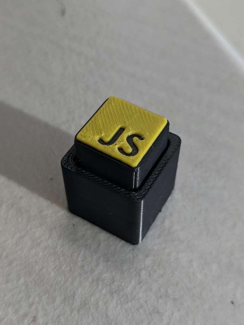 May include: A 3D-printed black and yellow square object. The top square is yellow with the letters "JS" cut out. The bottom square is black and slightly larger, creating a stepped effect. The object is likely a custom-made item.