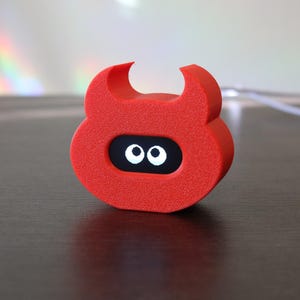 以下が含まれることがあります： 2 本の角と白い目の黒い楕円形の顔を持つ、明るい赤色の悪魔の形をしたオブジェクト。オブジェクトは濃い茶色の表面に置かれています。背景には白いコードが見えます。