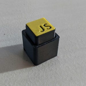 May include: A black, two-tiered, rectangular object with a yellow top. The top layer features the initials "JS" in a sans-serif font. The object appears to be 3D-printed.