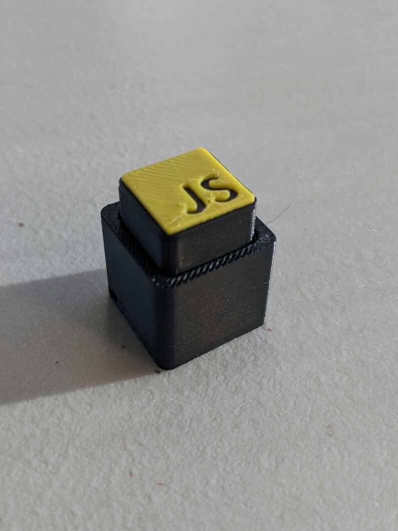 May include: A black, two-tiered, rectangular object with a yellow top. The top layer features the initials "JS" in a sans-serif font. The object appears to be 3D-printed.