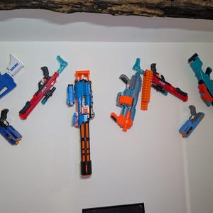 Puede incluir: Colección de blásters de juguete en varios colores, incluyendo azul, rojo y naranja, montados en una pared blanca. Los blásters varían en tamaño y diseño, algunos con accesorios adicionales.