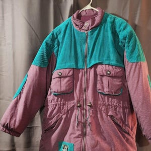 Op de afbeelding: Een vintage jas in tinten van teal en mauve. Het bovenste gedeelte en de schouderaccenten zijn van teal corduroy, terwijl de body mauve is. Voorzien van een ritssluiting, meerdere zakken en een taille met trekkoord. Een patch met een cartoonfiguur staat linksonder.