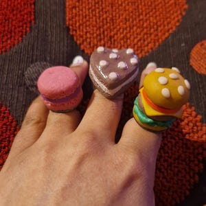 Könnte beinhalten: Drei ausgefallene Ringe: ein rosa Macaron, ein herzförmiger Kuchen und ein Miniatur-Hamburger. Der Macaron-Ring ist rosa mit einem hellblauen Sockel. Der Kuchenring ist braun mit weißen Punkten. Der Hamburger-Ring hat ein gelbes Brötchen, Käse und grünen Salat.