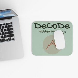 May include: A computer mouse and mousepad with the text "DeCoDe hidden mgs". The mousepad is light green with a beige circle and a red mountain graphic. A white mouse is on the mousepad, next to a laptop.
