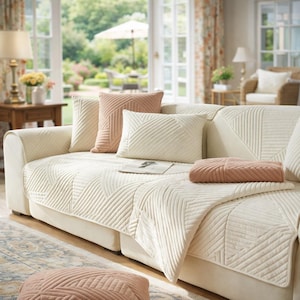 以下が含まれることがあります： クリーム色のソファに、いくつかの装飾用クッションとスローケットが置かれています。クッションとスローはクリーム色とダスティーローズの色合いです。ソファは幾何学模様のキルティング生地で覆われています。お揃いのオットマンが手前にあります。