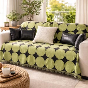 Op de afbeelding: Een bank met een groen en zwart patroon plaid. De plaid heeft grote, limoengroene cirkels op een zwarte achtergrond. Zwarte leren kussens en een crèmekleurig kussen met textuur liggen op de bank. De plaid heeft een gefranjerde rand.