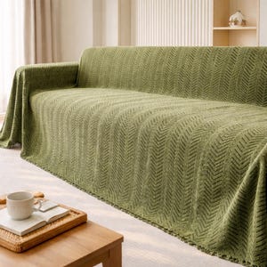 Puede incluir: Una funda de sofá verde con un patrón de chevron texturizado. La funda está colocada sobre un sofá, con los bordes colgando. Una pequeña mesa de madera con una bandeja, una taza y un libro están en primer plano.