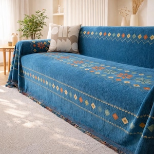 May include: A blue sofa cover with a geometric pattern in orange, yellow, and green. The cover has a fringed edge and is draped over a sofa. A decorative pillow rests on the sofa.