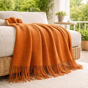以下が含まれることがあります： 白いソファに掛けられた、フリンジ付きのオレンジ色のテクスチャードスローブランケット。 ブランケットは織り模様で、柔らかい素材で作られています。 ソファは背景に緑のあるポーチにあります。 ブランケットの幅は約152cmです。