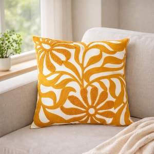 Op de afbeelding: Vierkantig sierkussen met een witte achtergrond en een groot, geborduurd bloemmotief in geeltinten. Het kussen ligt op een lichtgrijze bank, met een neutraal gekleurde deken in de buurt.