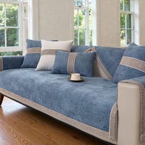 以下が含まれることがあります： 青いソファと、それに合うクッションとカバー。ソファはテクスチャード加工された生地で、ベージュのトリムが施されています。青とベージュの色合いのクッションがいくつかソファに配置されています。小さなカップとソーサーがソファの上にあります。