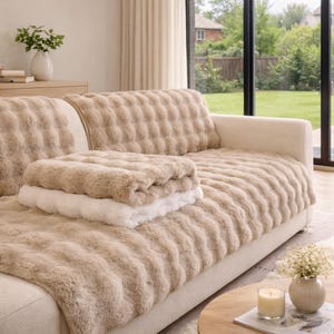 Könnte beinhalten: Ein beiges Sofa mit einem strukturierten, weichen Bezug und passendem Rückenkissenbezug. Eine gefaltete Decke in Beige- und Weißtönen liegt auf dem Sofa. Der Raum hat eine neutrale Farbpalette mit natürlichem Licht.