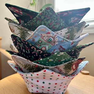 May include: A stack of five fabric bowl cozies featuring various holiday-themed patterns, including gingerbread houses, candy canes, holly leaves, and Christmas trees. The bottom cozy has a white base with red and green polka dots.