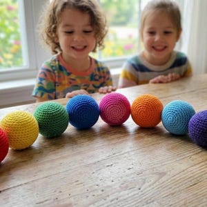 May include: A collection of colorful, crocheted balls in various colors, including red, yellow, green, blue, pink, orange, and purple, are arranged on a wooden table. Two children are smiling in the background. A decorative vase is also visible.