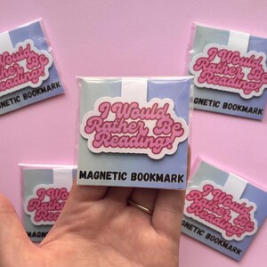 May include: Magnetic bookmarks with the text "I Would Rather Be Reading!" in pink and white, packaged in clear plastic. The bookmarks have a light blue and white background. The words "MAGNETIC BOOKMARK" are printed in black.