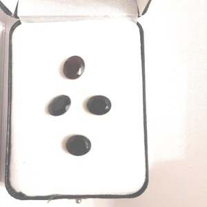 May include: A black jewellery box with a white interior holds four oval-shaped gemstones. One gemstone is a dark red colour, while the other three are black. The gemstones are displayed against a white background.