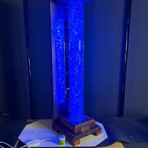 May include: A tall, illuminated decorative column with a wooden frame. The column features two clear, rectangular tubes filled with blue, sparkling material. The wooden base and top are stained a dark brown. A power cord and adapter are visible.