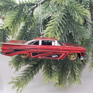 May include: A red die-cast toy car ornament featuring black graphic side detailing, gold-coloured wheels, and a silver-toned roof trim. The vehicle is suspended by a thin silver string, showcasing a classic car body style with a Hot Wheels logo.