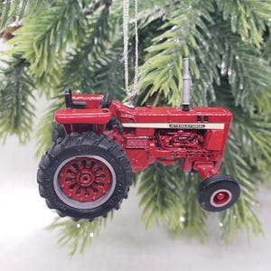 May include: A miniature red International tractor ornament featuring large black rear tires with red rims, a small black front tire, a vertical exhaust pipe, and a black seat. The word INTERNATIONAL is printed in white on the side of the engine hood.