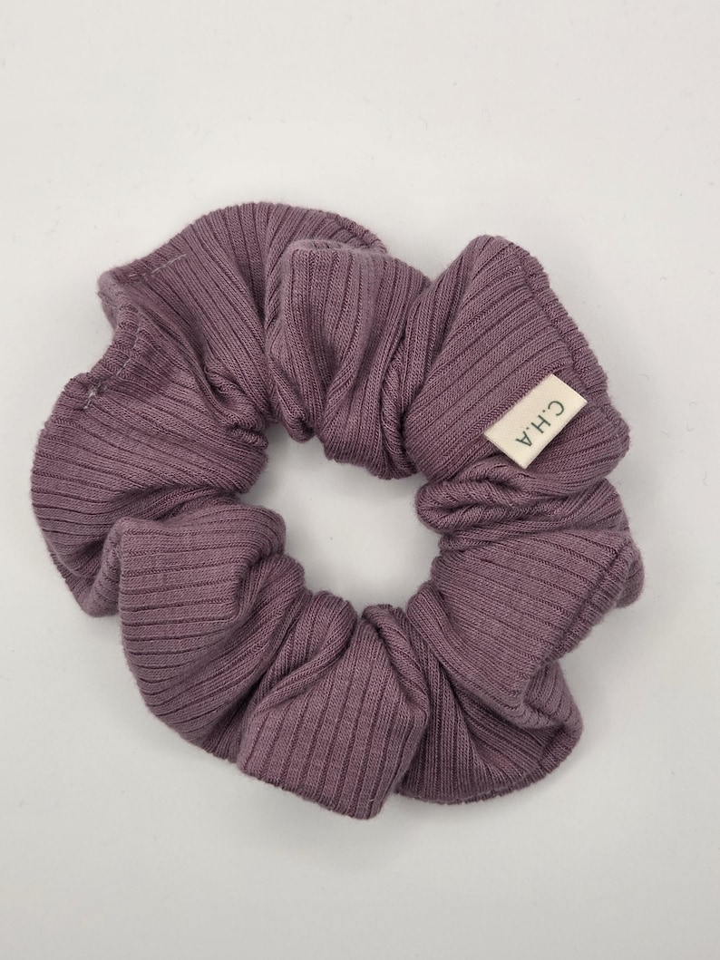 Puede incluir: Una goma para el pelo de color malva hecha de tela acanalada. Una peque&ntilde;a etiqueta rectangular con las letras "C.H.A" est&aacute; cosida en la goma. La goma es circular y parece estar hecha de un material suave.