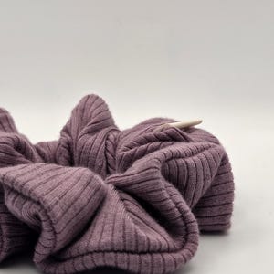 Puede incluir: Primer plano de una scrunchie de punto acanalado malva. La tela es suave y fruncida, con acanalado vertical. Un peque&ntilde;o palito de madera de color claro est&aacute; insertado en la scrunchie.