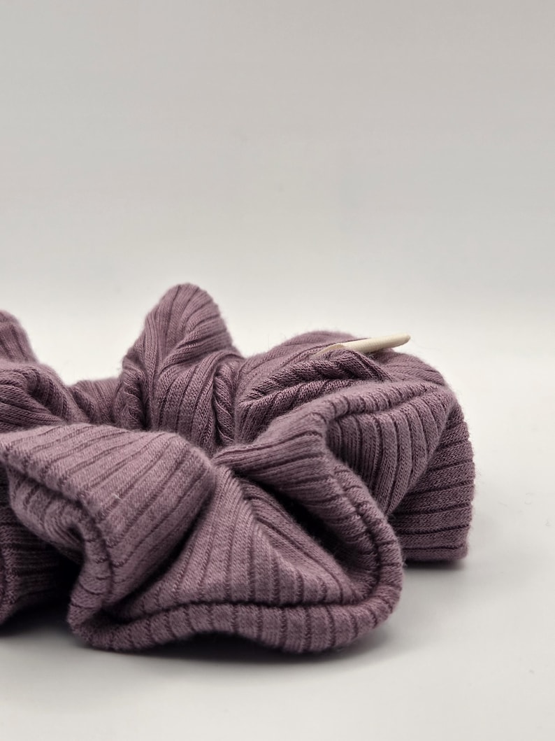 Puede incluir: Primer plano de una scrunchie de punto acanalado malva. La tela es suave y fruncida, con acanalado vertical. Un peque&ntilde;o palito de madera de color claro est&aacute; insertado en la scrunchie.