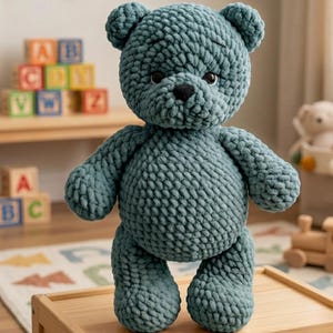 Könnte beinhalten: Ein handgefertigter, teal-farbener Häkelteddybär mit einer schwarzen Nase und kleinen schwarzen Augen. Der Plüschbär steht auf einer Holzoberfläche. Der Bär hat ein strukturiertes, grob gestricktes Aussehen mit sichtbaren Maschen. Der Hintergrund umfasst Holzblöcke und ein Spielzeug.