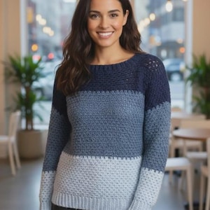 May include: A blue, grey, and light blue crochet sweater with a round neckline. The sweater has long sleeves and a textured stitch pattern. The woman in the image is smiling.