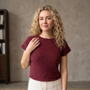 Puede incluir: Un top de ganchillo burdeos con mangas cortas. El top tiene un cuello acanalado y un patrón de puntada texturizada. La modelo lleva el top con pantalones blancos. El top es un artículo hecho a mano.