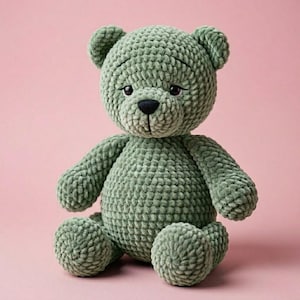 May include: A handmade, sage green crochet teddy bear with a black nose and small, dark eyes. The plush bear is sitting upright with its arms and legs extended. The background is a solid pink.