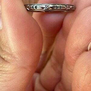 May include: A silver-colored ring with an engraved floral pattern. The ring is held between two fingers, showcasing the intricate design. The ring's band is thin, and the floral pattern is repeated around the entire circumference.