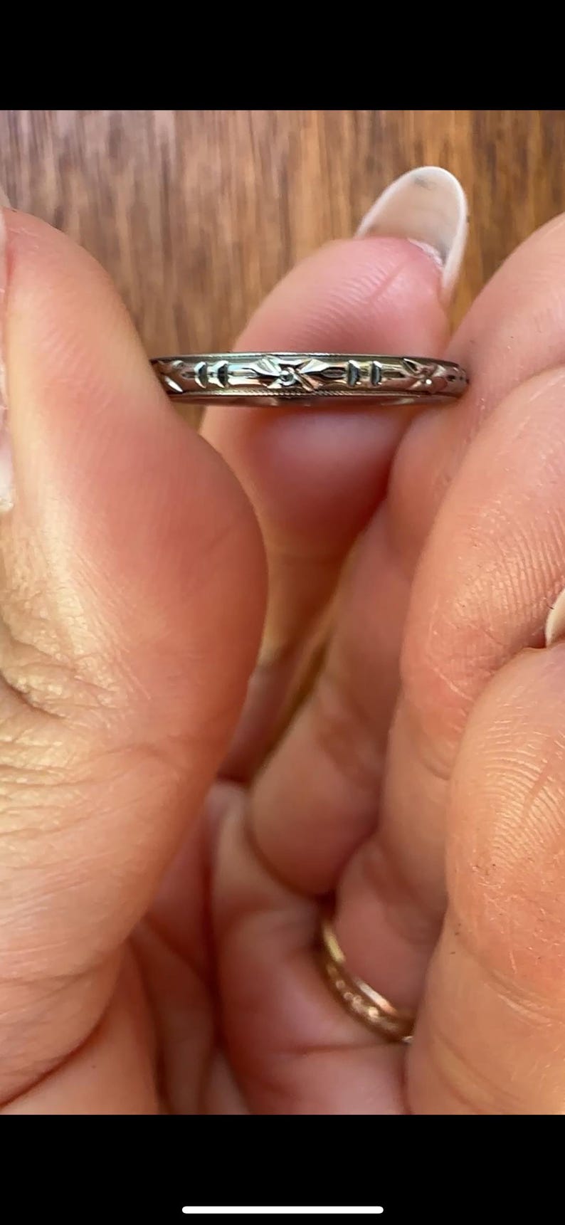 May include: A silver-colored ring with an engraved floral pattern. The ring is held between two fingers, showcasing the intricate design. The ring's band is thin, and the floral pattern is repeated around the entire circumference.
