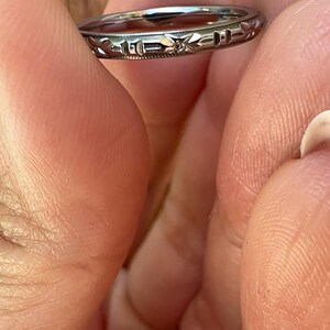May include: A silver ring with an engraved design featuring geometric shapes and floral elements. The ring is held between fingers, showcasing its intricate details and craftsmanship. The ring's design is visible, highlighting its vintage or antique style.