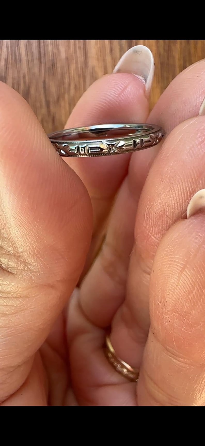 May include: A silver ring with an engraved design featuring geometric shapes and floral elements. The ring is held between fingers, showcasing its intricate details and craftsmanship. The ring's design is visible, highlighting its vintage or antique style.