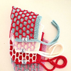 May include: A stack of knitted baby bonnets in various colors, including red, pink, and blue. Each bonnet features a honeycomb design and ties. The label "dover & madden" is visible on each bonnet.