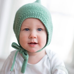 Peut inclure: Un bébé portant un bonnet en tricot vert avec un sommet pointu et une chemise blanche à manches longues. Le bonnet a un nœud noué à l'arrière.