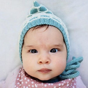 Puede incluir: Un bebé con un gorro de punto azul y blanco con la parte superior puntiaguda. El gorro tiene un patrón geométrico blanco y azul. El bebé lleva un babero de lunares rosa y blanco.