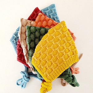 May include: A stack of six knitted baby bonnets in various colours, including yellow, green, orange, red, beige, and blue. The bonnets are made with a waffle stitch pattern and have a knotted tie.