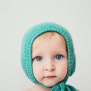 May include: A baby wearing a teal knitted hat with a matching scarf. The baby has blonde hair and blue eyes.