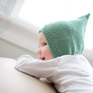 Può includere: Un bambino che indossa un berretto di lana verde chiaro con la punta a punta, sdraiato su una superficie marrone chiaro e guarda a destra.