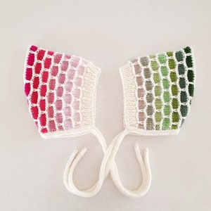 May include: Two knitted baby bonnets with a honeycomb pattern. One bonnet is pink, red, and white, and the other is green and white.