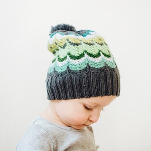 Puede incluir: Un niño con un gorro de punto gris y verde con un pompón en la parte superior. El gorro tiene un patrón ondulado en tonos de verde y blanco.
