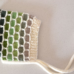 May include: A knitted baby hat with a green and white geometric pattern and white ties.