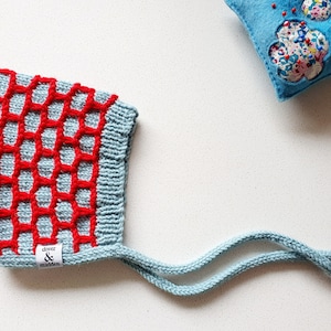 May include: A light blue knitted baby bonnet with a red honeycomb pattern. The bonnet has a ribbed edge and ties, and a small white tag with the text "dover & madden". A blue felt flower-shaped pin cushion is in the background.