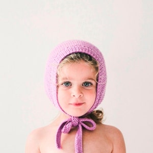 以下が含まれることがあります： 紫色のニットのボンネットとそれに合わせたリボンをつけた小さな子供。ボンネットはシンプルなデザインで、柔らかな糸で作られています。