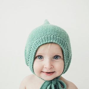 May include: A baby wearing a light green knitted bonnet with a pointed top. The baby is looking directly at the camera and smiling.