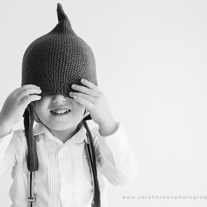 以下が含まれることがあります： 目はグレーのニット帽で覆われている子供。帽子は尖った先端と長い紐がついている。子供は白い襟付きシャツを着ている。