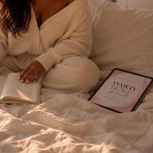 Può includere: Una persona con un completo in maglia color crema, che legge un libro e usa un tablet. Il tablet mostra il testo "Anxiety Dump Journal" in un design rosa e bianco. La scena è ambientata su un letto con biancheria da letto color crema.