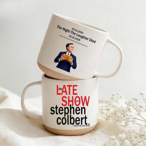 May include: Two white ceramic mugs stacked on top of each other. The top mug features an image of a man holding a sandwich and the text "The Night The Laughter Died." The bottom mug has "The Late Show with Stephen Colbert" printed on it.