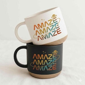 Puede incluir: Dos tazas de café de cerámica, una blanca y otra negra, apiladas. Cada taza presenta la palabra "AMAZE" en un diseño de gradiente de arcoíris, con un gráfico de planeta. Las tazas tienen una base marrón y un asa blanca.